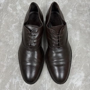 May include: A pair of dark brown leather oxford shoes featuring a cap toe design, a closed lacing system with thin matching laces, and a smooth polished finish. The shoes have a rounded toe shape and a low heel, showcasing a clean and structured silhouette.
