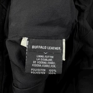 May include: A black garment label featuring white text that reads BUFFALO LEATHER, followed by a list of lining translations and the composition of 100% polyester. The label is stitched into the interior of a black fabric garment with a smooth, dark lining.