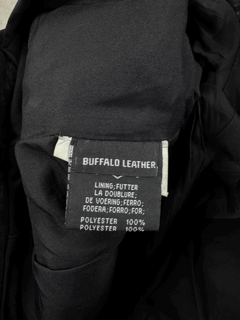 May include: A black garment label featuring white text that reads BUFFALO LEATHER, followed by a list of lining translations and the composition of 100% polyester. The label is stitched into the interior of a black fabric garment with a smooth, dark lining.