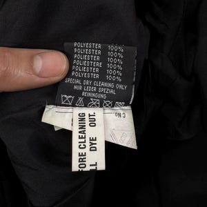 May include: A black garment care label featuring the text POLYESTER 100% in multiple languages, SPECIAL DRY CLEANING ONLY, and NUR LEDER SPEZIAL REINIGUNG. A secondary white tag attached below reads BEFORE CLEANING ALL DYE OUT and C No.