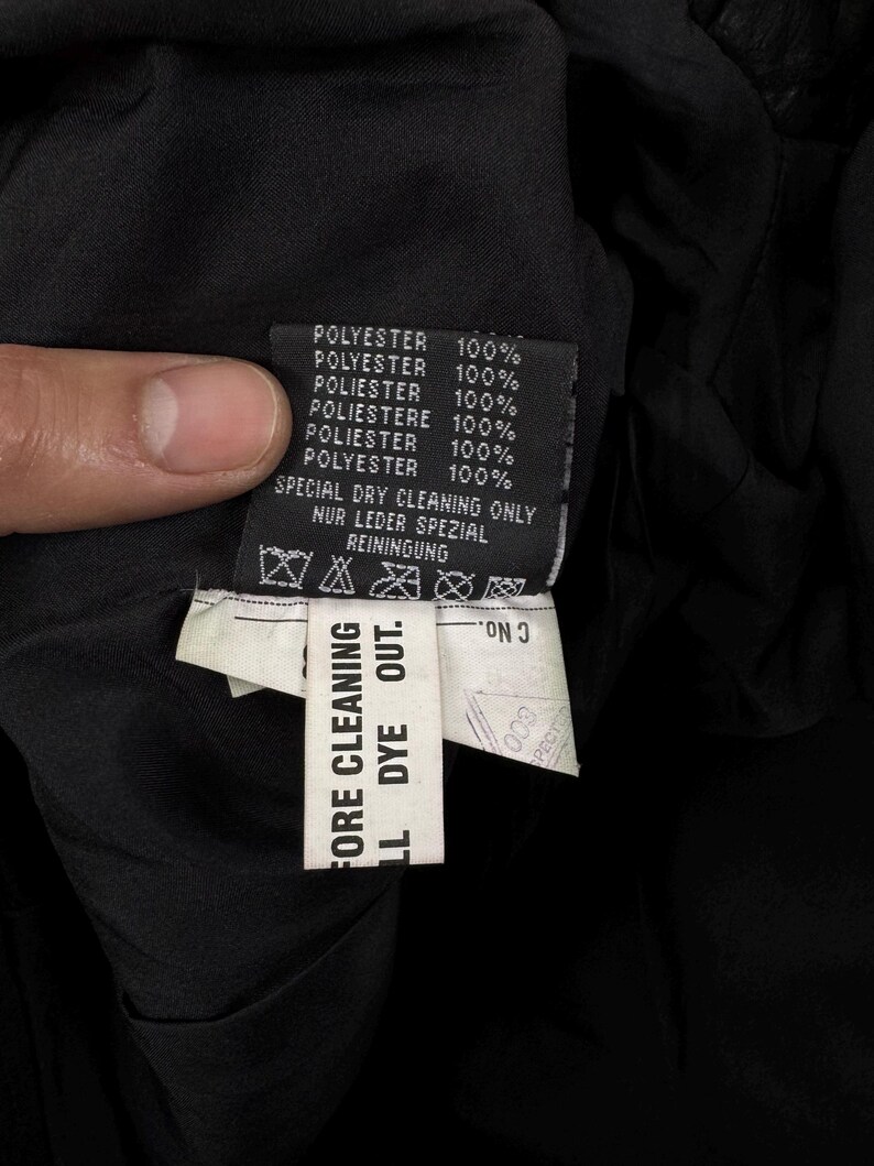 May include: A black garment care label featuring the text POLYESTER 100% in multiple languages, SPECIAL DRY CLEANING ONLY, and NUR LEDER SPEZIAL REINIGUNG. A secondary white tag attached below reads BEFORE CLEANING ALL DYE OUT and C No.