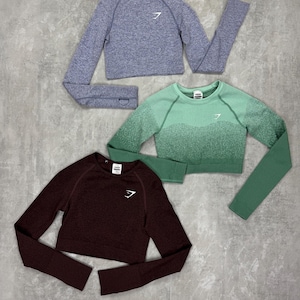 May include: Three cropped long-sleeve shirts in shades of blue, green, and burgundy. Each shirt features a crew neck and a white logo on the chest. The shirts are made of a textured material.