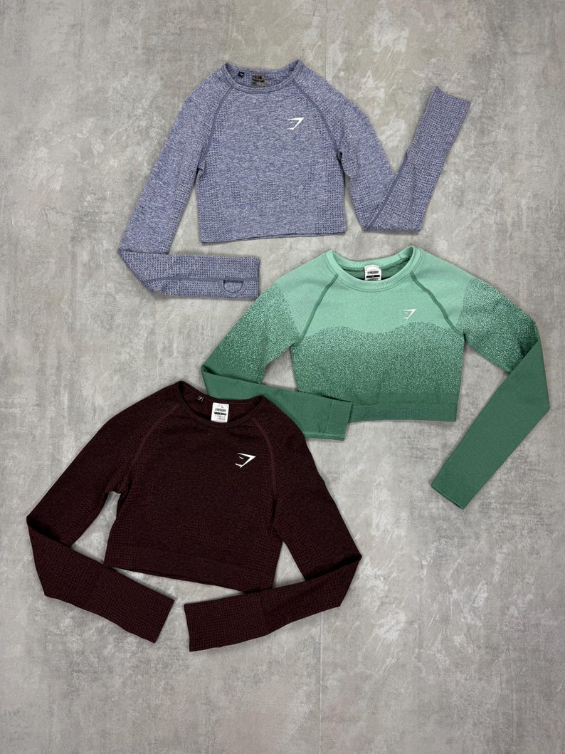May include: Three cropped long-sleeve shirts in shades of blue, green, and burgundy. Each shirt features a crew neck and a white logo on the chest. The shirts are made of a textured material.
