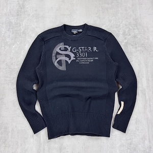 May include: Black long-sleeve sweater with a ribbed texture. The sweater features a large embroidered graphic with the text "G-STAR R. 3301 SOUTH-EAST DISTRICT 5304 SPEC SUPPORT SYSTEM G-STAR RAW".