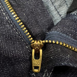 Puede incluir: Una vista de cerca de una tela de mezclilla azul &iacute;ndigo oscuro con un cierre de cremallera de metal color lat&oacute;n. El tirador de la cremallera tiene grabadas las letras YKK y los dientes de la cremallera tienen un acabado en tono lat&oacute;n a juego.