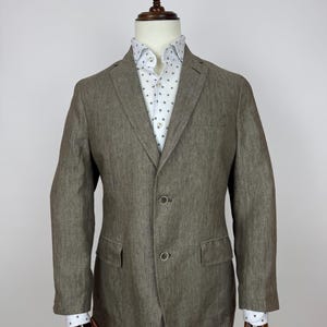 May include: A brown blazer with two buttons and two front pockets, worn over a white shirt with a small, repeating pattern. The blazer is displayed on a mannequin.
