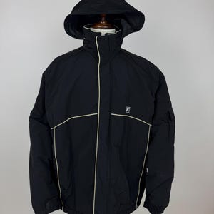 Puede incluir: Chaqueta negra con capucha, ribete blanco y el logo de Fila. La chaqueta tiene una cremallera completa, un cuello alto y una capucha. La chaqueta est&aacute; hecha de un material duradero y resistente al agua. La chaqueta se muestra en un maniqu&iacute;.