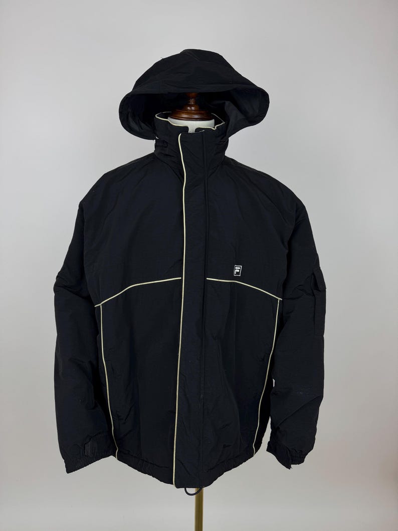 Puede incluir: Chaqueta negra con capucha, ribete blanco y el logo de Fila. La chaqueta tiene una cremallera completa, un cuello alto y una capucha. La chaqueta est&aacute; hecha de un material duradero y resistente al agua. La chaqueta se muestra en un maniqu&iacute;.