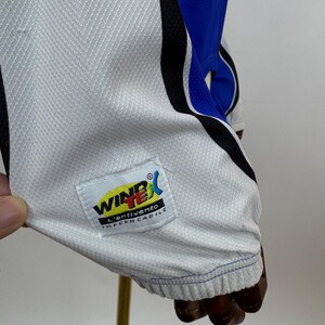 May include: Close-up of a white and blue athletic garment. The garment features a white mesh fabric with blue and black accents. A label with the text "WIND X" and "L'ANTIVENTO IMPERMEABILE" is visible.