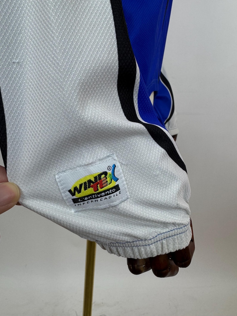 May include: Close-up of a white and blue athletic garment. The garment features a white mesh fabric with blue and black accents. A label with the text "WIND X" and "L'ANTIVENTO IMPERMEABILE" is visible.