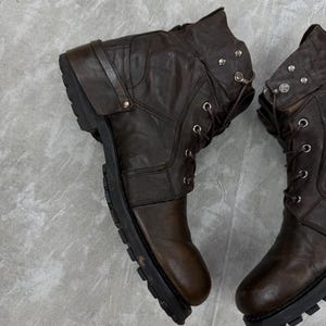 May include: A pair of brown leather boots with a rugged, worn appearance. The boots feature a lace-up design, metal accents, and a thick, black sole. The boots have a folded-over top and a strap detail.