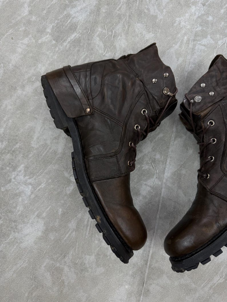 May include: A pair of brown leather boots with a rugged, worn appearance. The boots feature a lace-up design, metal accents, and a thick, black sole. The boots have a folded-over top and a strap detail.