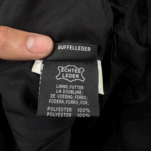 May include: A black garment care label featuring the text BUFFELLEDER, ECHTES LEDER, and a list of translations for lining including FUTTER, LA DOUBLURE, DE VOERING, FERRO, FODERA, FORRO, and FOR, followed by the specification POLYESTER 100%.