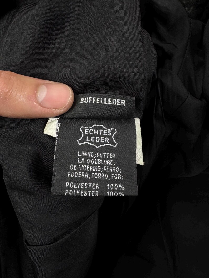 May include: A black garment care label featuring the text BUFFELLEDER, ECHTES LEDER, and a list of translations for lining including FUTTER, LA DOUBLURE, DE VOERING, FERRO, FODERA, FORRO, and FOR, followed by the specification POLYESTER 100%.