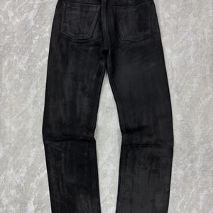 May include: A pair of black trousers featuring a textured, matte finish with a straight-leg cut. The trousers include a standard waistband with belt loops, a button closure, and two patch pockets on the back. The fabric displays subtle tonal variations.