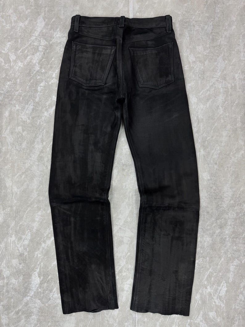 May include: A pair of black trousers featuring a textured, matte finish with a straight-leg cut. The trousers include a standard waistband with belt loops, a button closure, and two patch pockets on the back. The fabric displays subtle tonal variations.
