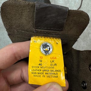 May include: Close-up of a brown leather boot with a yellow tag. The tag indicates a USA size 13, UK size 12, and EUR size 46. The boot is made with leather upper and man-made materials.