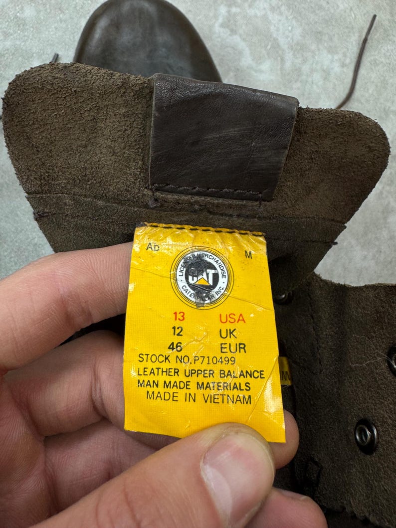 May include: Close-up of a brown leather boot with a yellow tag. The tag indicates a USA size 13, UK size 12, and EUR size 46. The boot is made with leather upper and man-made materials.