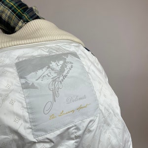 Puede incluir: Chaqueta blanca con un parche blanco en la espalda que dice "Montecore Dolomiti The Luxury Sport". El parche tiene un dise&ntilde;o de cordillera.