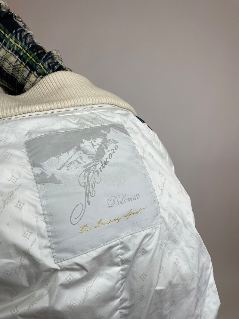 Puede incluir: Chaqueta blanca con un parche blanco en la espalda que dice "Montecore Dolomiti The Luxury Sport". El parche tiene un dise&ntilde;o de cordillera.