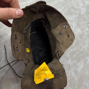 May include: Brown leather work boot with a folded-over cuff. The boot has metal eyelets and laces. A yellow tag with text is visible inside the boot. The brand name "Caterpillar" is visible inside the boot.