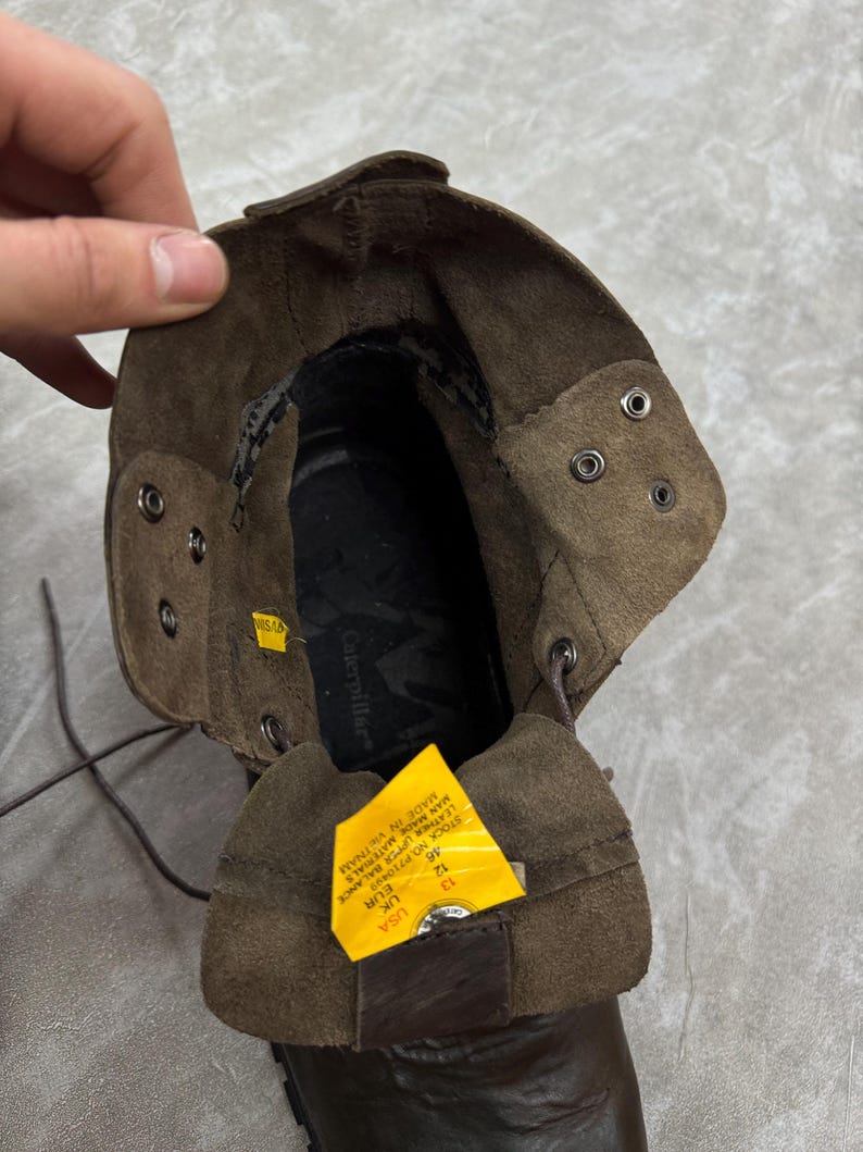 May include: Brown leather work boot with a folded-over cuff. The boot has metal eyelets and laces. A yellow tag with text is visible inside the boot. The brand name "Caterpillar" is visible inside the boot.