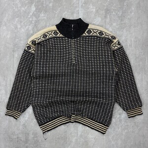 Puede incluir: Un suéter negro y crema con cremallera y un patrón geométrico. El suéter tiene un cuello alto, una cremallera de media longitud y detalles en crema en los hombros y los puños. El dobladillo y los puños tienen rayas crema y negras.