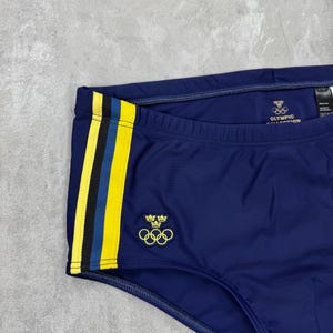 May include: Navy blue swim trunks with yellow and blue side stripes. The trunks feature the Olympic rings logo and the text "OLYMPIC COLLECTION". A small emblem with crowns and rings is also visible.
