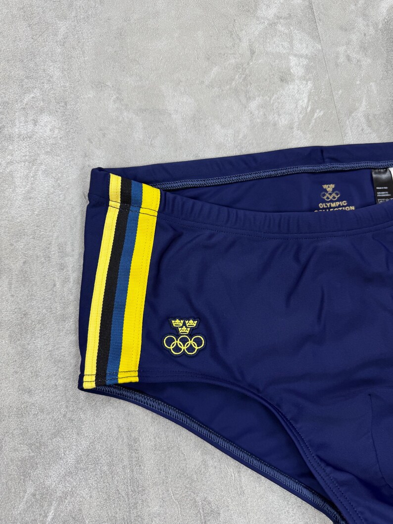 May include: Navy blue swim trunks with yellow and blue side stripes. The trunks feature the Olympic rings logo and the text "OLYMPIC COLLECTION". A small emblem with crowns and rings is also visible.