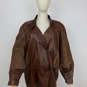 Puede incluir: Chaqueta de cuero marrón con diseño cruzado. La chaqueta presenta un cuello ancho, dos bolsillos delanteros y una mezcla de paneles de cuero liso y de ante. La chaqueta se exhibe en un maniquí.