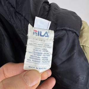 Puede incluir: Primer plano de una etiqueta de chaqueta Fila. La etiqueta es blanca con el logotipo de Fila en rojo y azul. El texto de la etiqueta incluye "100% Poliammide Nylon" y "Made in Macau."