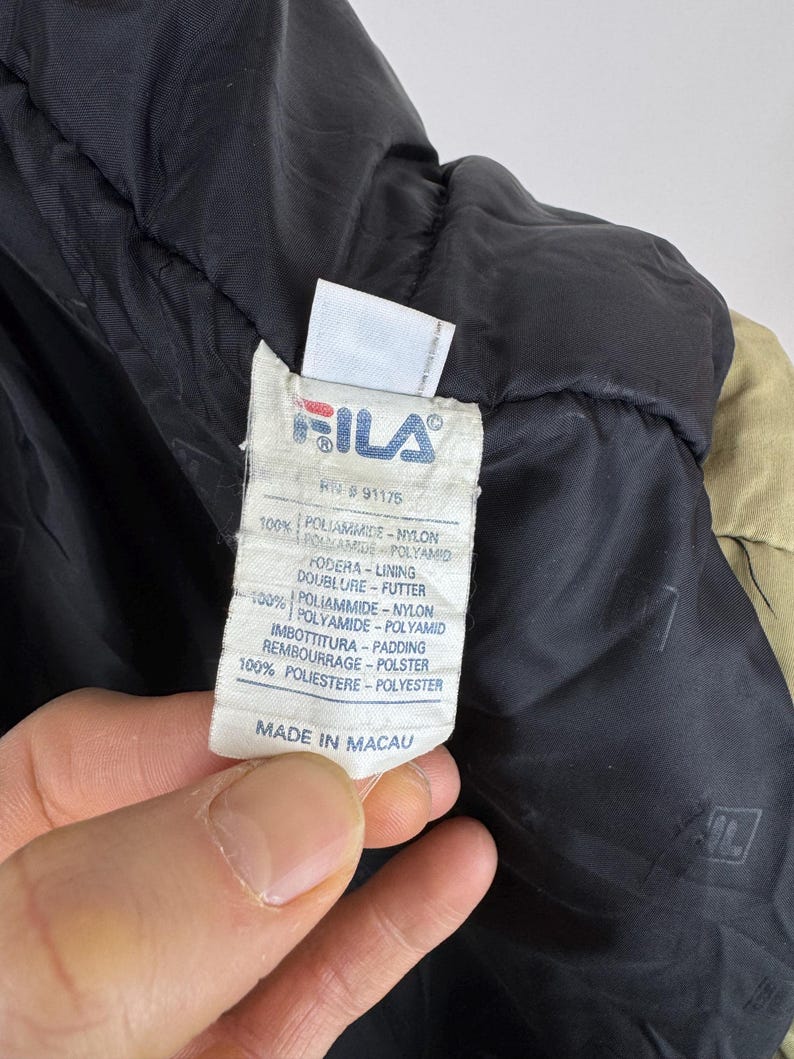 Puede incluir: Primer plano de una etiqueta de chaqueta Fila. La etiqueta es blanca con el logotipo de Fila en rojo y azul. El texto de la etiqueta incluye "100% Poliammide Nylon" y "Made in Macau."