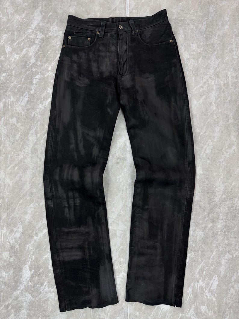 May include: Black denim jeans featuring a distressed, washed-out aesthetic with lighter grey streaks throughout the fabric. These trousers include a standard button fly closure, five pockets, and a straight-leg cut with a clean, finished hemline.
