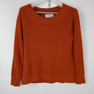 Könnte beinhalten: Ein rostfarbener Pullover mit einem breiten, gerippten Ausschnitt und langen Ärmeln. Der Pullover ist aus einem strukturierten Strickstoff gefertigt und hängt an einem weißen Kleiderbügel. Das Etikett lautet "Collection Avenue".