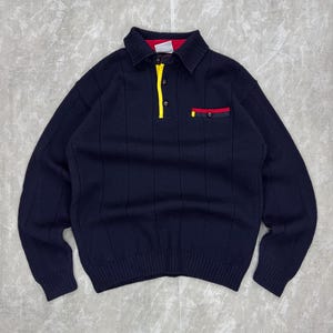 May include: Navy blue long-sleeve sweater with a collared neckline. The sweater features a yellow stripe down the placket and a pocket with red and yellow accents. The ribbed knit design adds texture to the garment.