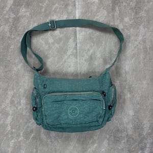 Op de afbeelding: Teal crossbody tas met meerdere vakken met ritssluiting en een lange verstelbare riem. De tas heeft een cirkelvormig logo op de voorkant en zilverkleurige hardware. De tas is gemaakt van een textuurrijk materiaal.