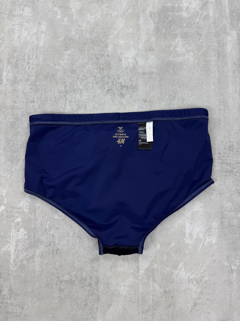 May include: Navy blue swim trunks with a tag. The trunks have a high waist and a dark blue color. The tag reads "Olympic Collection" and "H&M".