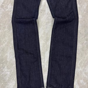 Puede incluir: Un par de pantalones vaqueros de mezclilla azul &iacute;ndigo oscuro con piernas rectas y costuras blancas en contraste a lo largo de las costuras exteriores e interiores. La tela tiene una textura de sarga visible y un corte recto y estructurado.