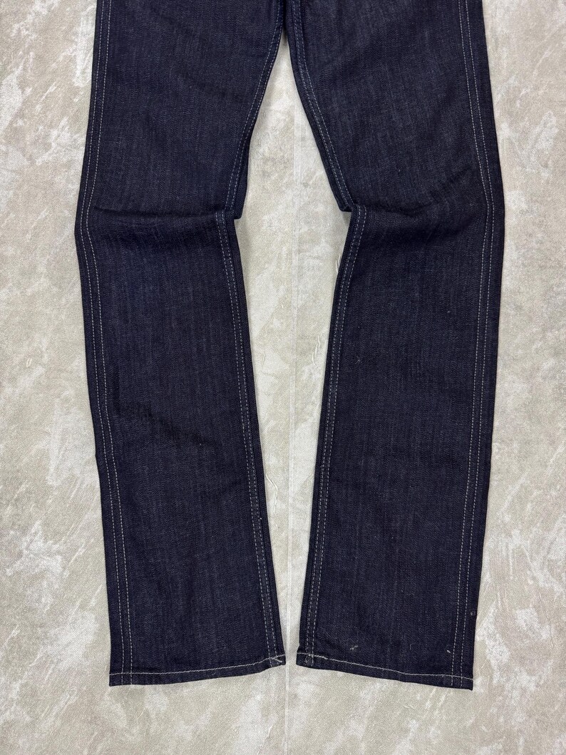 Puede incluir: Un par de pantalones vaqueros de mezclilla azul &iacute;ndigo oscuro con piernas rectas y costuras blancas en contraste a lo largo de las costuras exteriores e interiores. La tela tiene una textura de sarga visible y un corte recto y estructurado.