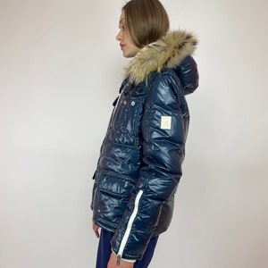 Puede incluir: Una chaqueta acolchada azul marino con capucha de piel sint&eacute;tica y detalles de cremallera blanca. La chaqueta tiene un acabado brillante y un dise&ntilde;o acolchado.
