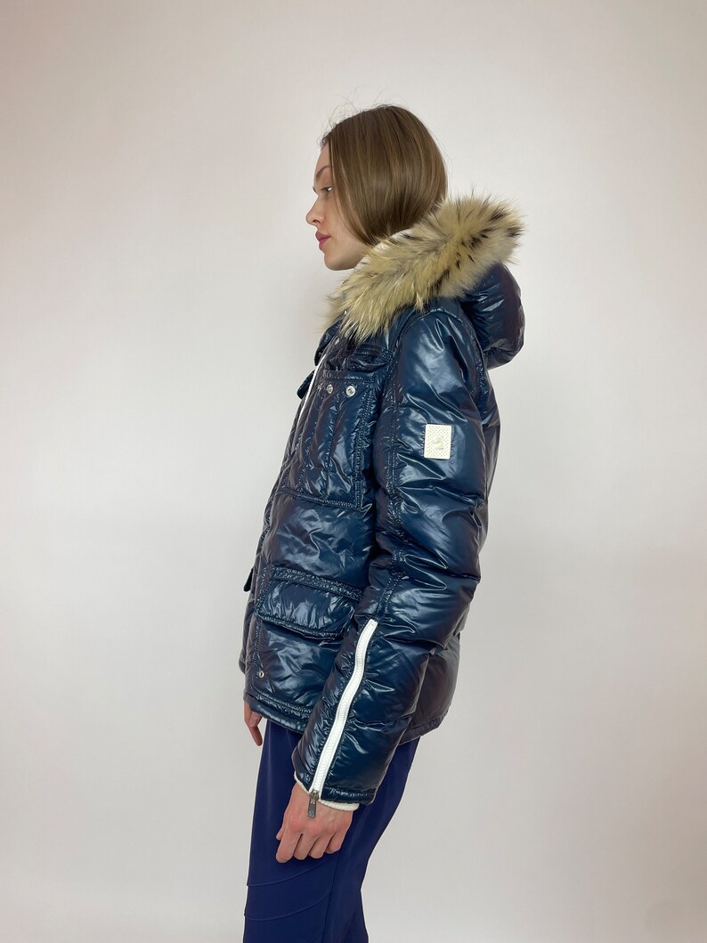 Puede incluir: Una chaqueta acolchada azul marino con capucha de piel sint&eacute;tica y detalles de cremallera blanca. La chaqueta tiene un acabado brillante y un dise&ntilde;o acolchado.