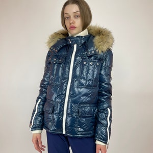 Puede incluir: Una chaqueta acolchada azul con una cremallera blanca y ribetes blancos. La chaqueta tiene una capucha de piel sint&eacute;tica y varios bolsillos.