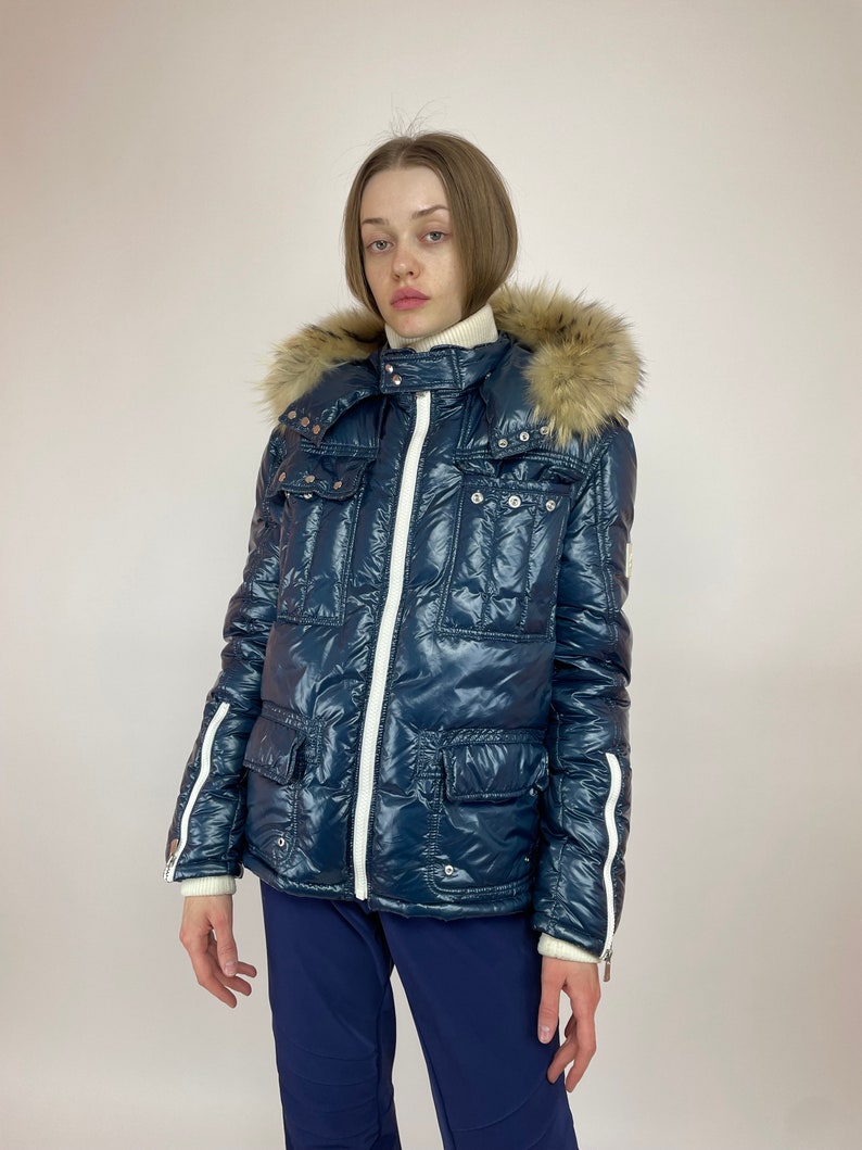 Puede incluir: Una chaqueta acolchada azul con una cremallera blanca y ribetes blancos. La chaqueta tiene una capucha de piel sint&eacute;tica y varios bolsillos.
