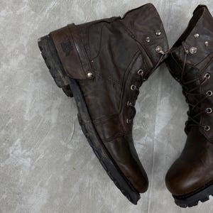 May include: Pair of worn brown leather boots with laces and metal eyelets. The boots have a rugged, distressed appearance, with visible wear and tear on the leather and soles. The boots are designed for durability and style.