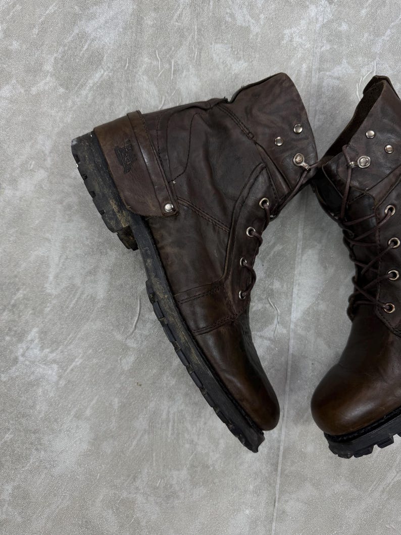 May include: Pair of worn brown leather boots with laces and metal eyelets. The boots have a rugged, distressed appearance, with visible wear and tear on the leather and soles. The boots are designed for durability and style.