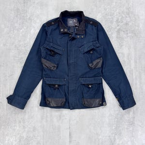 May include: A navy blue utility jacket with multiple pockets and a black collar. The jacket has a zipper closure and button accents.