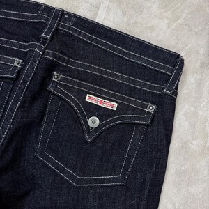 Puede incluir: Vaqueros de mezclilla azul &iacute;ndigo oscuro con un bolsillo trasero con solapa en punta, un bot&oacute;n plateado y una etiqueta rectangular con el texto Hudson Jeans y un peque&ntilde;o gr&aacute;fico de la bandera Union Jack. Los vaqueros tienen costuras blancas.