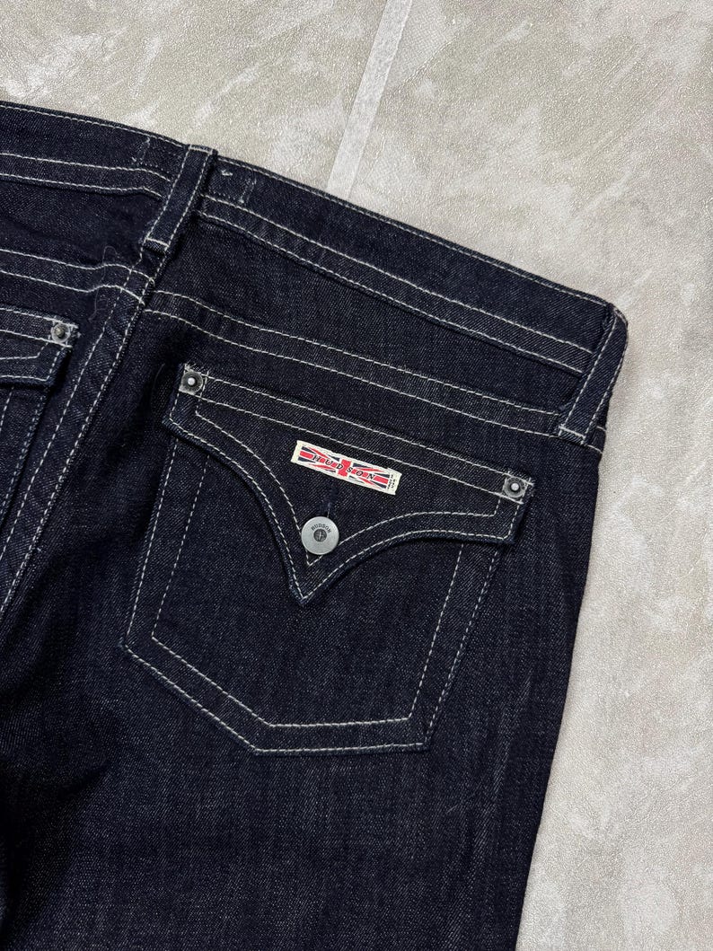 Puede incluir: Vaqueros de mezclilla azul &iacute;ndigo oscuro con un bolsillo trasero con solapa en punta, un bot&oacute;n plateado y una etiqueta rectangular con el texto Hudson Jeans y un peque&ntilde;o gr&aacute;fico de la bandera Union Jack. Los vaqueros tienen costuras blancas.