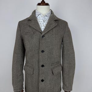 Puede incluir: Chaqueta marrón con estampado de espiga, solapas con muescas, tres botones negros y dos bolsillos delanteros. La chaqueta se exhibe en un maniquí y se combina con una camisa blanca con un pequeño estampado azul.