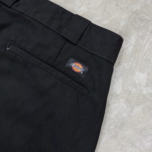 May include: Black work trousers featuring a structured waistband with belt loops and a single rear welt pocket. A rectangular Dickies brand logo patch with red, yellow, and blue text is stitched above the pocket on the right side of the garment.